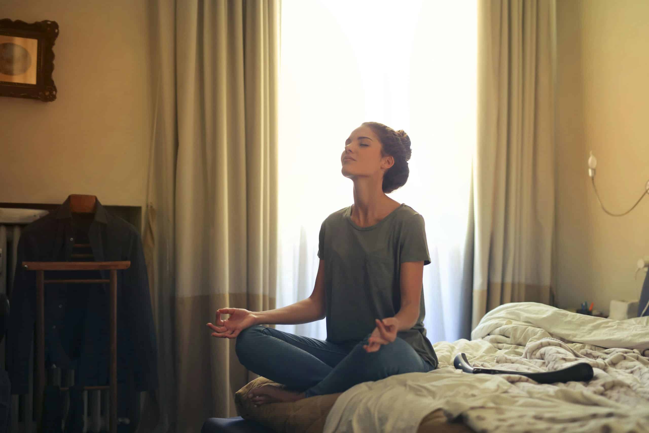 A woman in jeans and a t-shirt meditating on her bed.